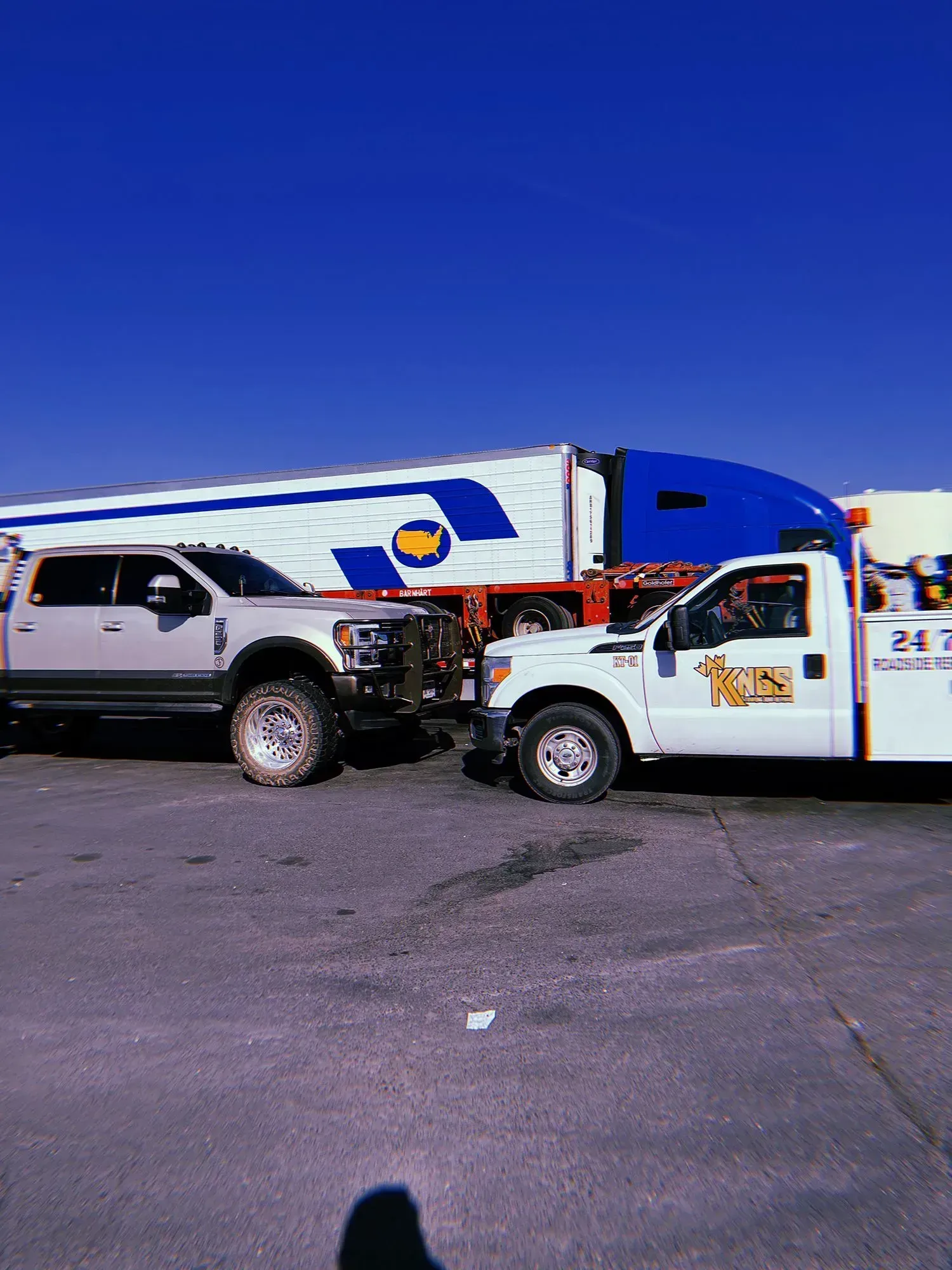 A white service truck and a silver pickup truck parked in front of a blue and white semi-trailer on an asphalt lot.