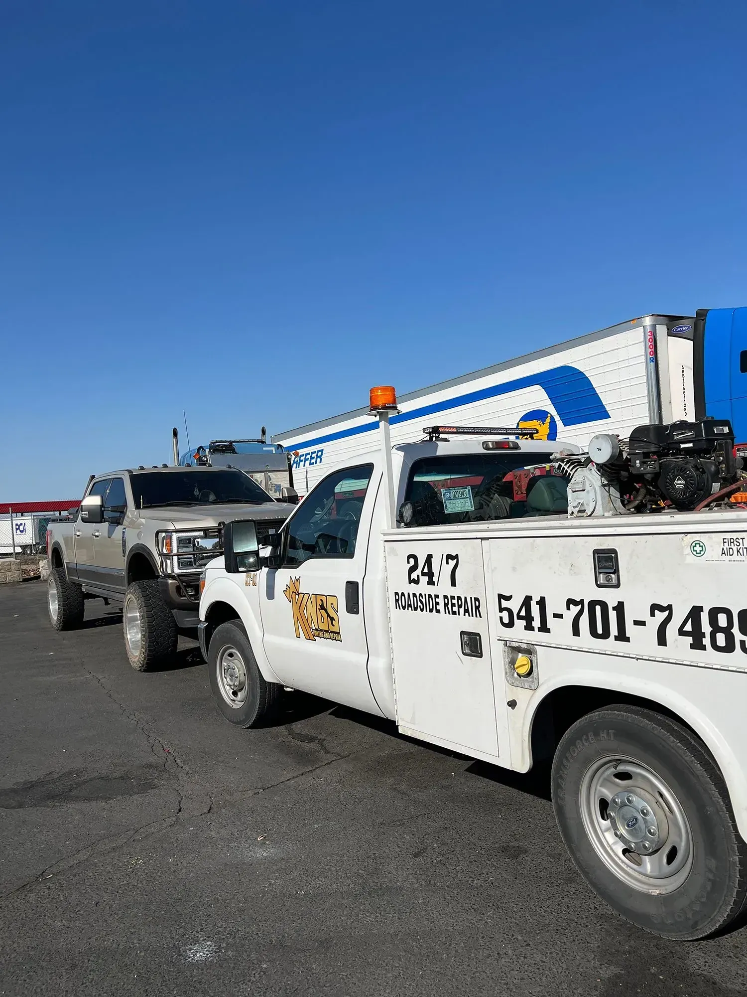 A white roadside service truck parked next to a lifted silver pickup truck against a bright blue sky.