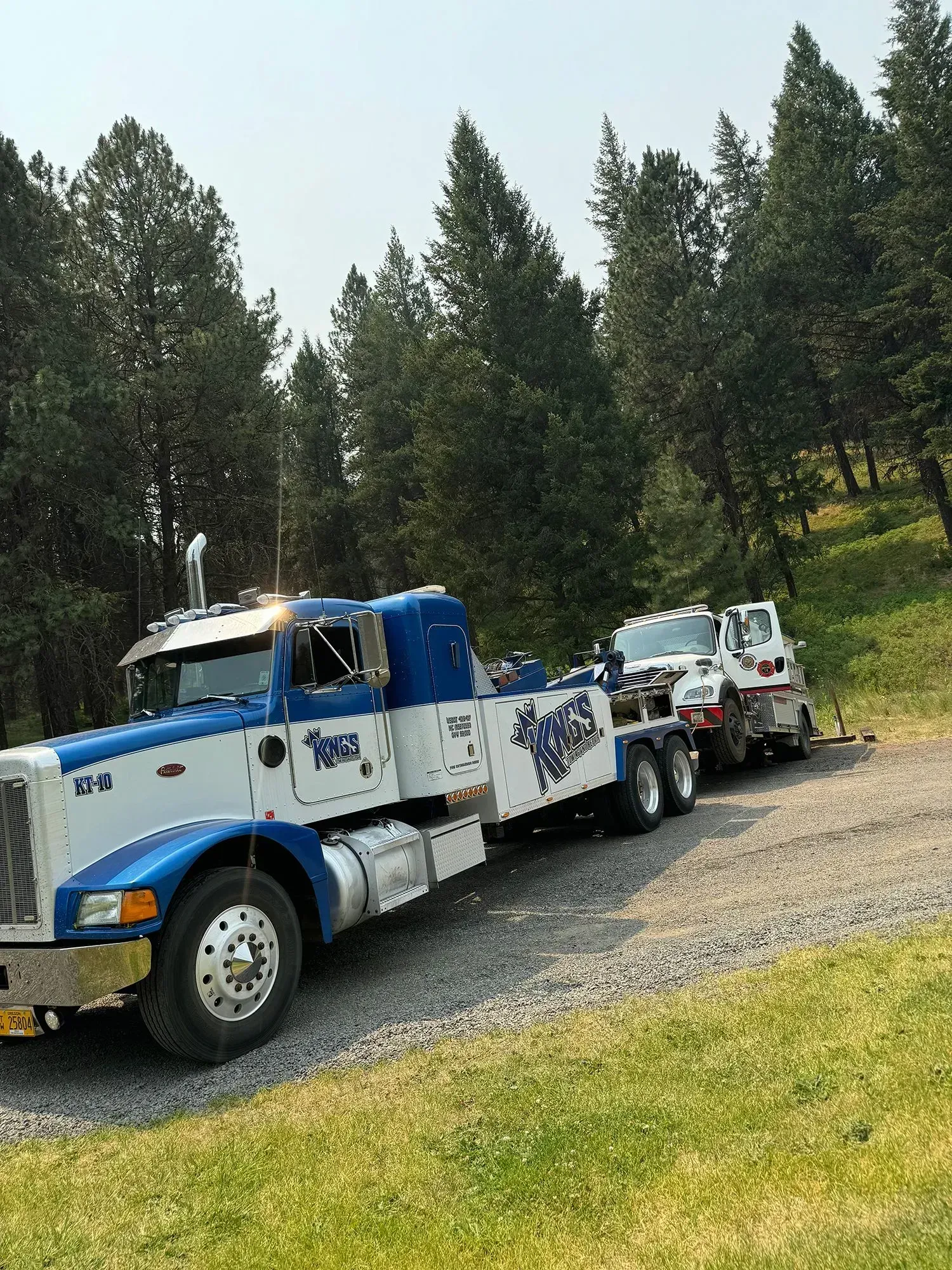 A blue and white heavy-duty tow truck is parked on a gravel shoulder next to a forest, towing a smaller white truck.