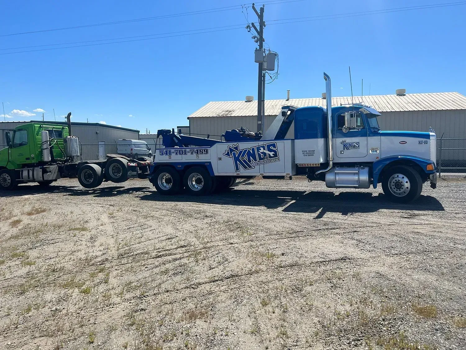 A white and blue tow truck tows a green semi-truck across a gravel lot on a sunny day.