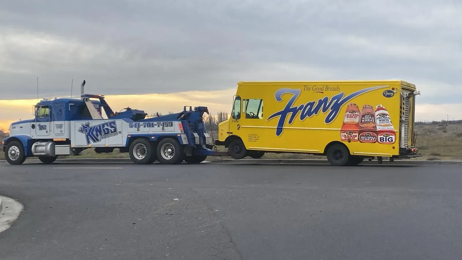 A blue tow truck pulling a yellow Franz delivery van along a road under an overcast sky.