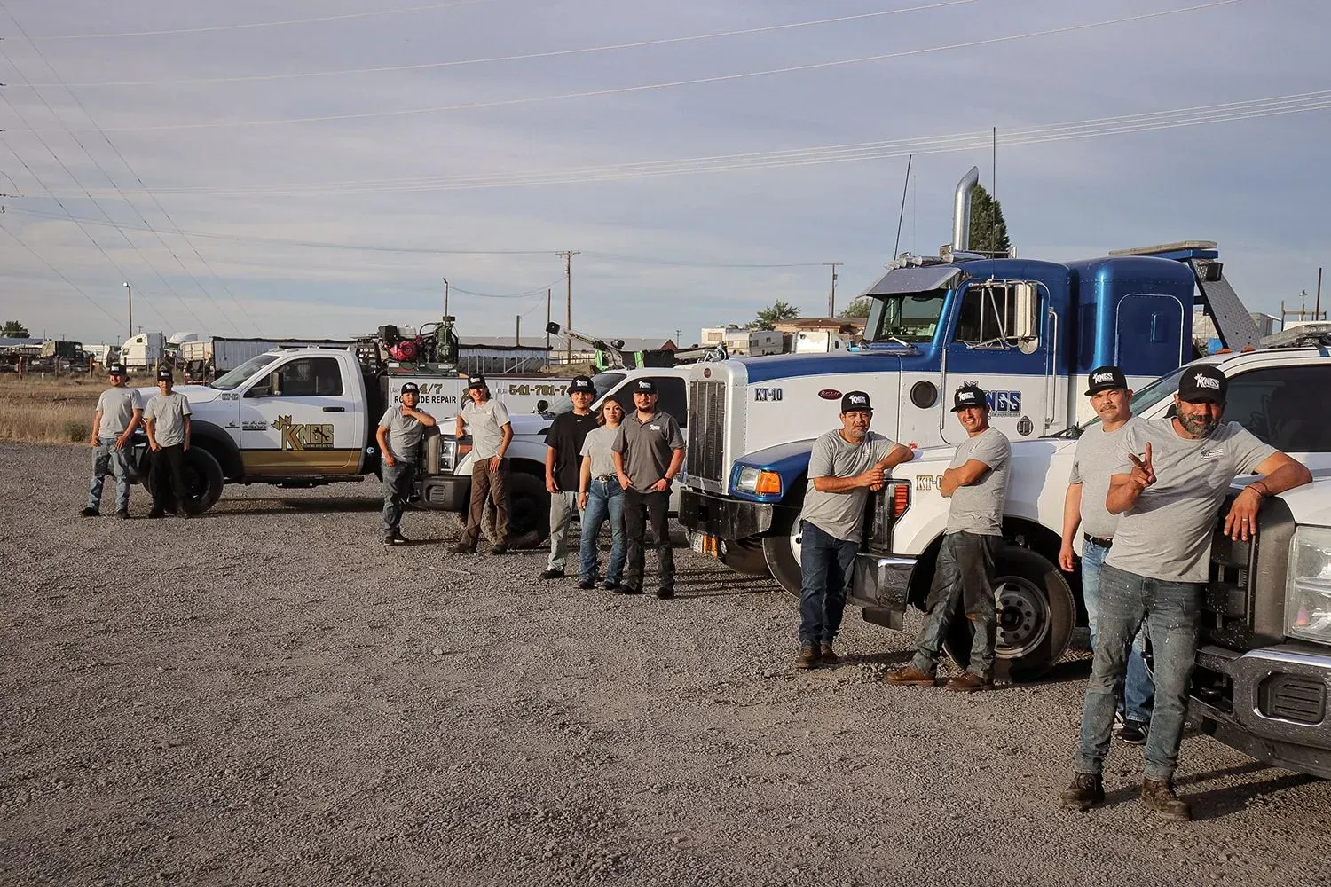A group of people wearing matching gray T-shirts and caps standing in a gravel lot with several tow trucks.