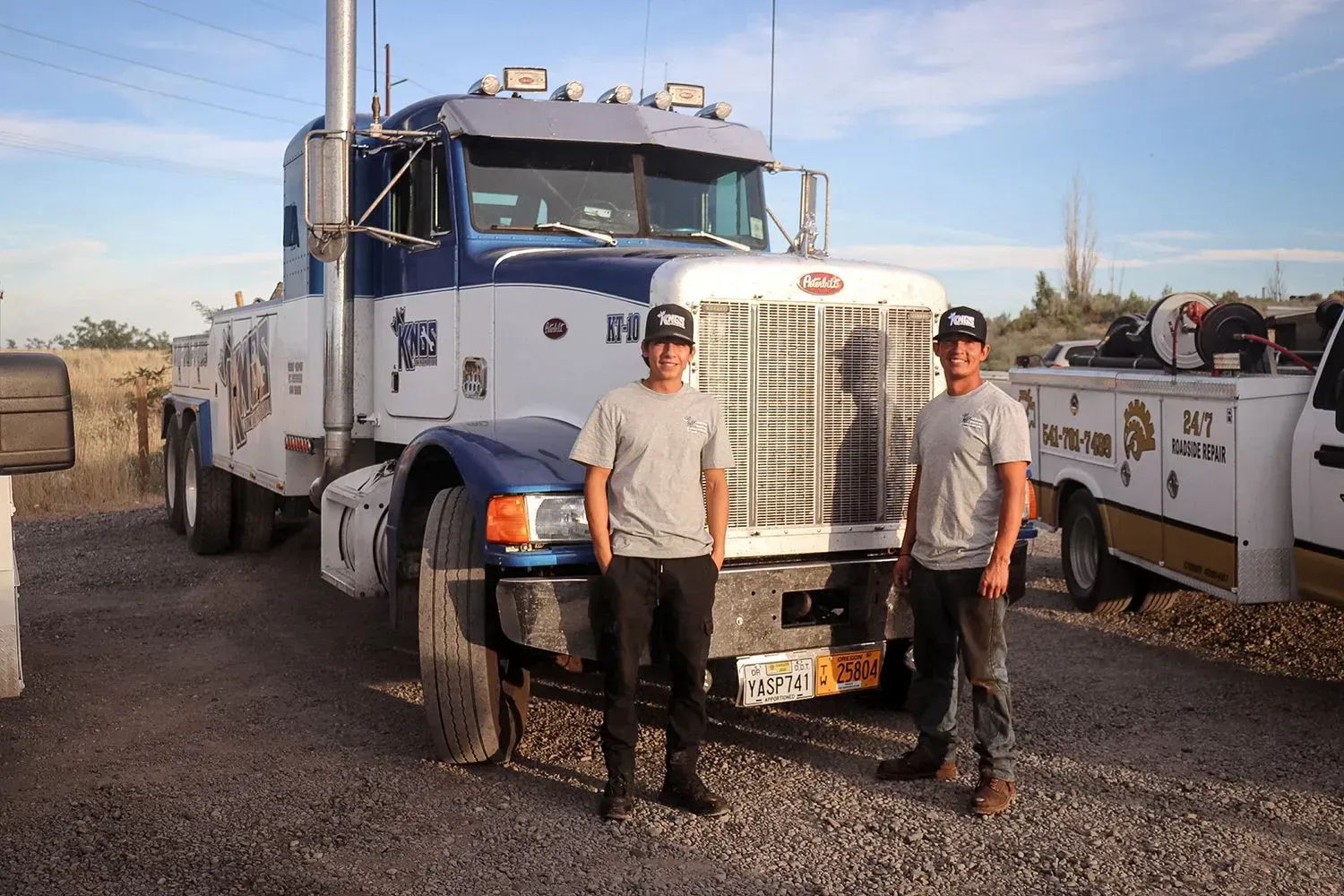 Two people stand in front of a blue and white tow truck parked on a gravel lot during the day.