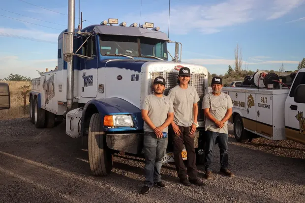 Three people stand in front of a blue and white tow truck and a service truck parked on a gravel lot under a blue sky.