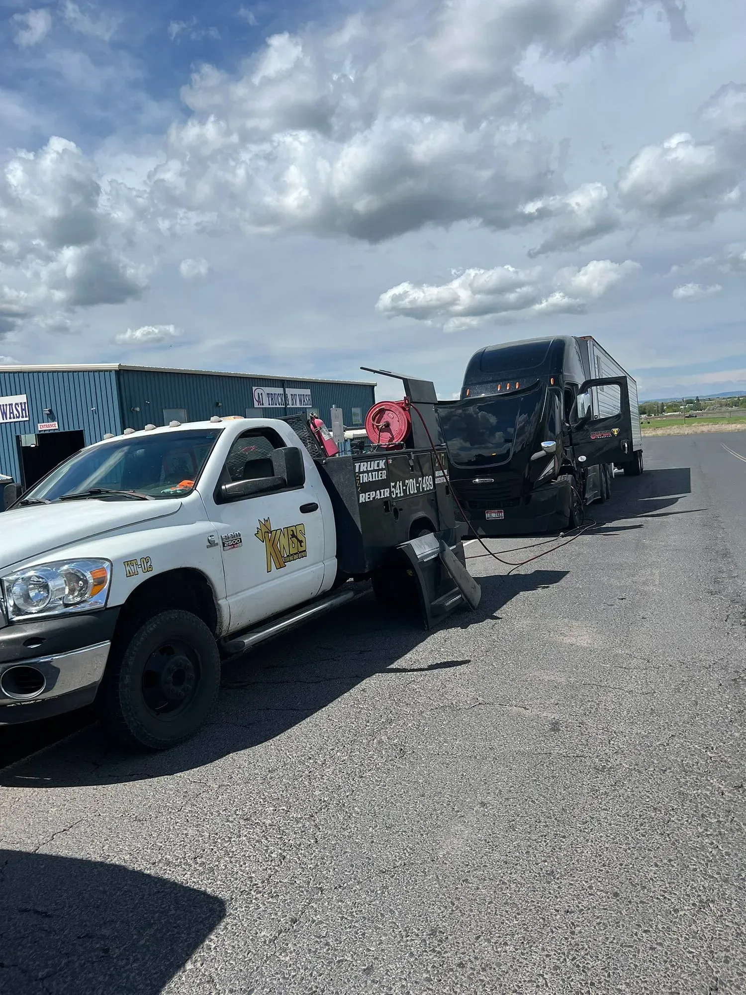 A white service truck towing a black semi-truck cab on a paved lot under a cloudy sky.