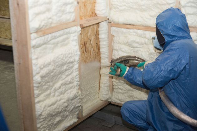 A person in protective blue gear uses a spray nozzle to apply white insulating foam to a wooden wall frame.
