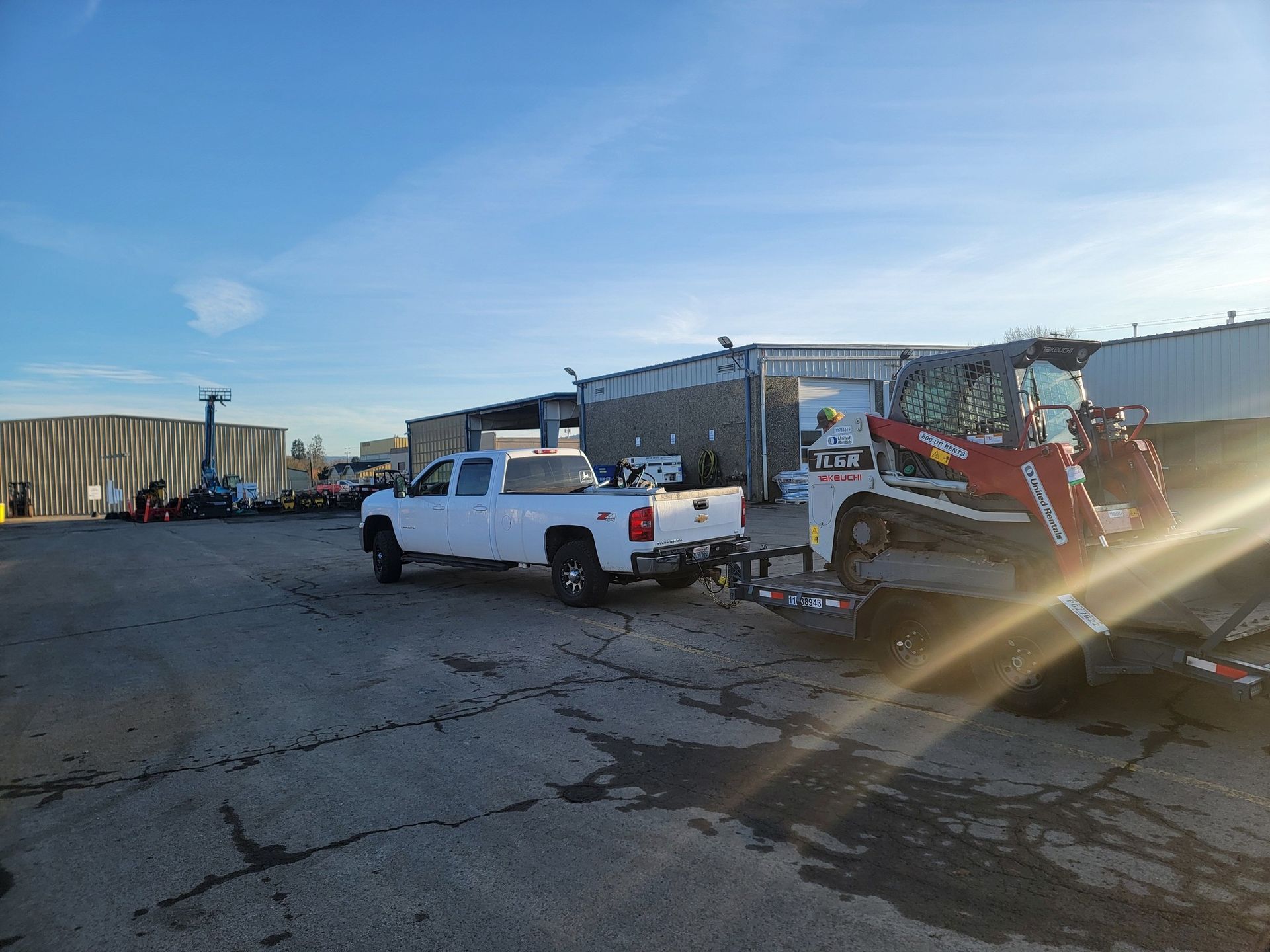 A white truck is towing a forklift in a parking lot.