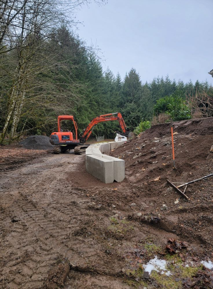 An excavator is driving down a dirt road next to a concrete wall.