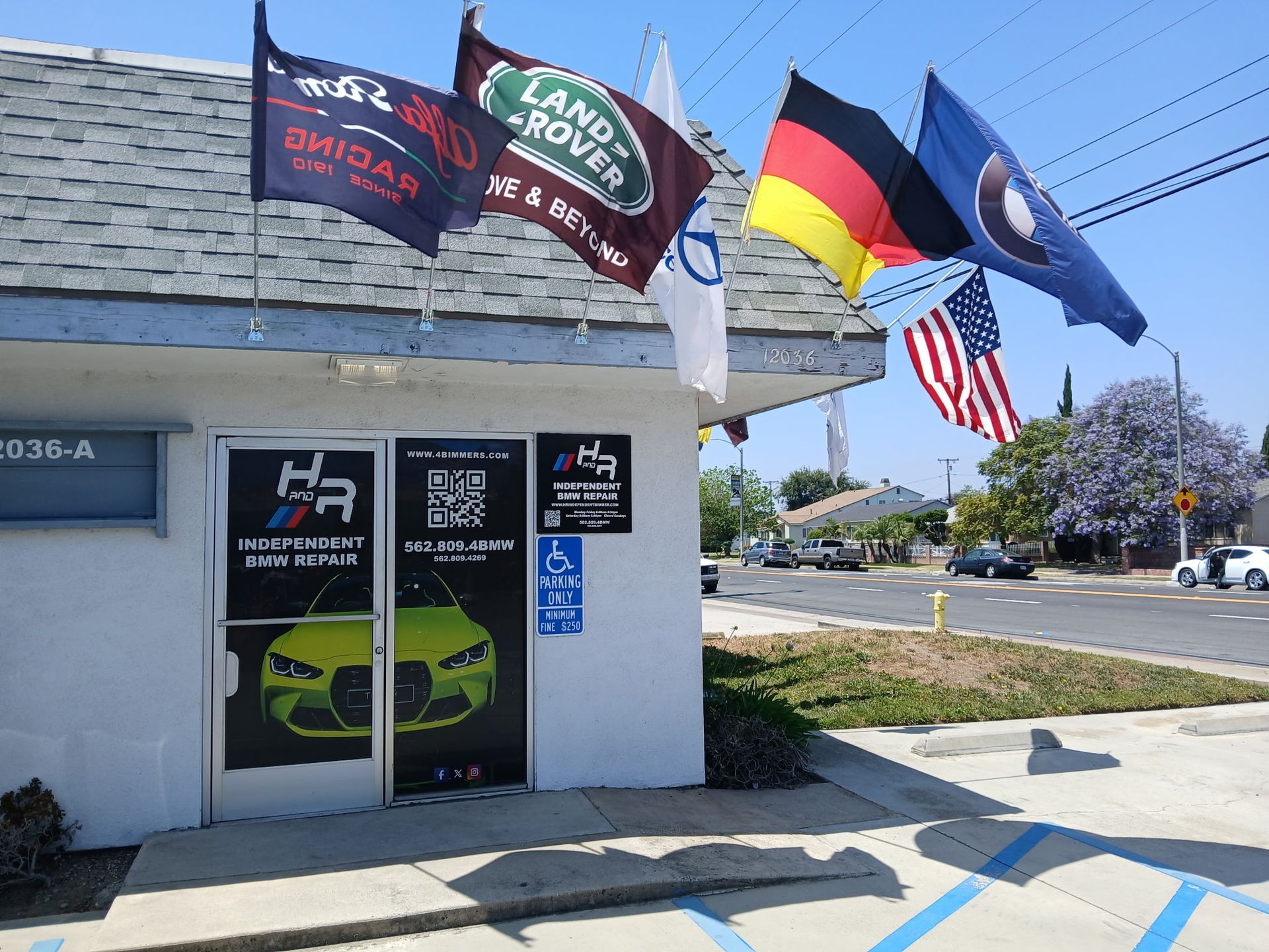 Auto shop with a green car graphic on the door, flanked by multiple flags outside on a sunny street.