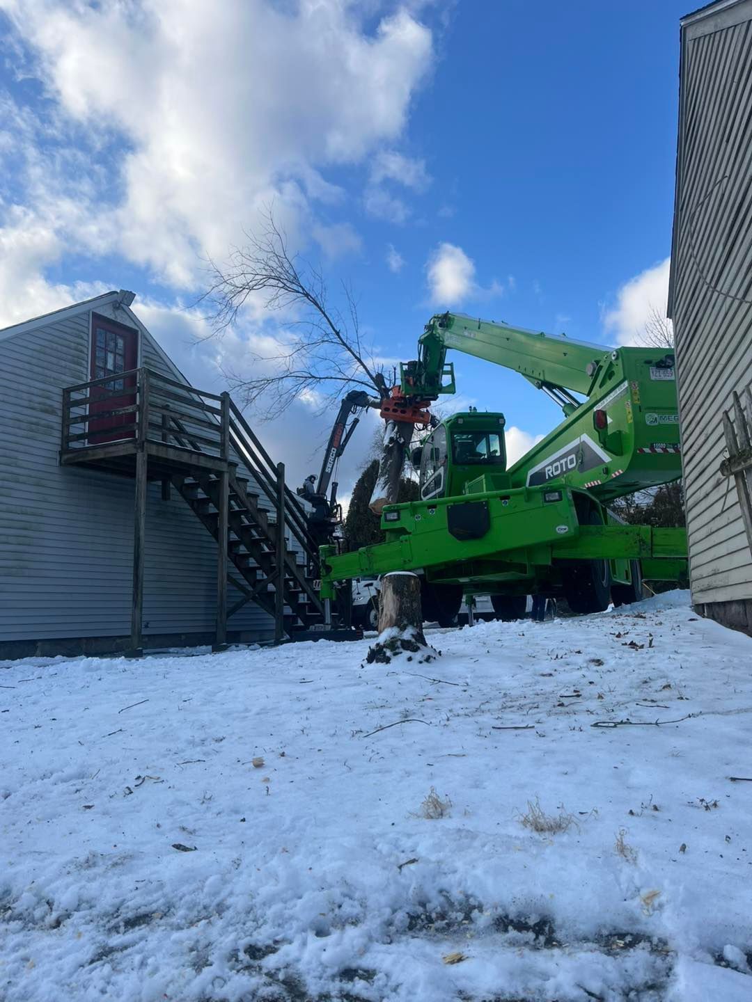 A green crane is cutting a tree in the snow in front of a house.