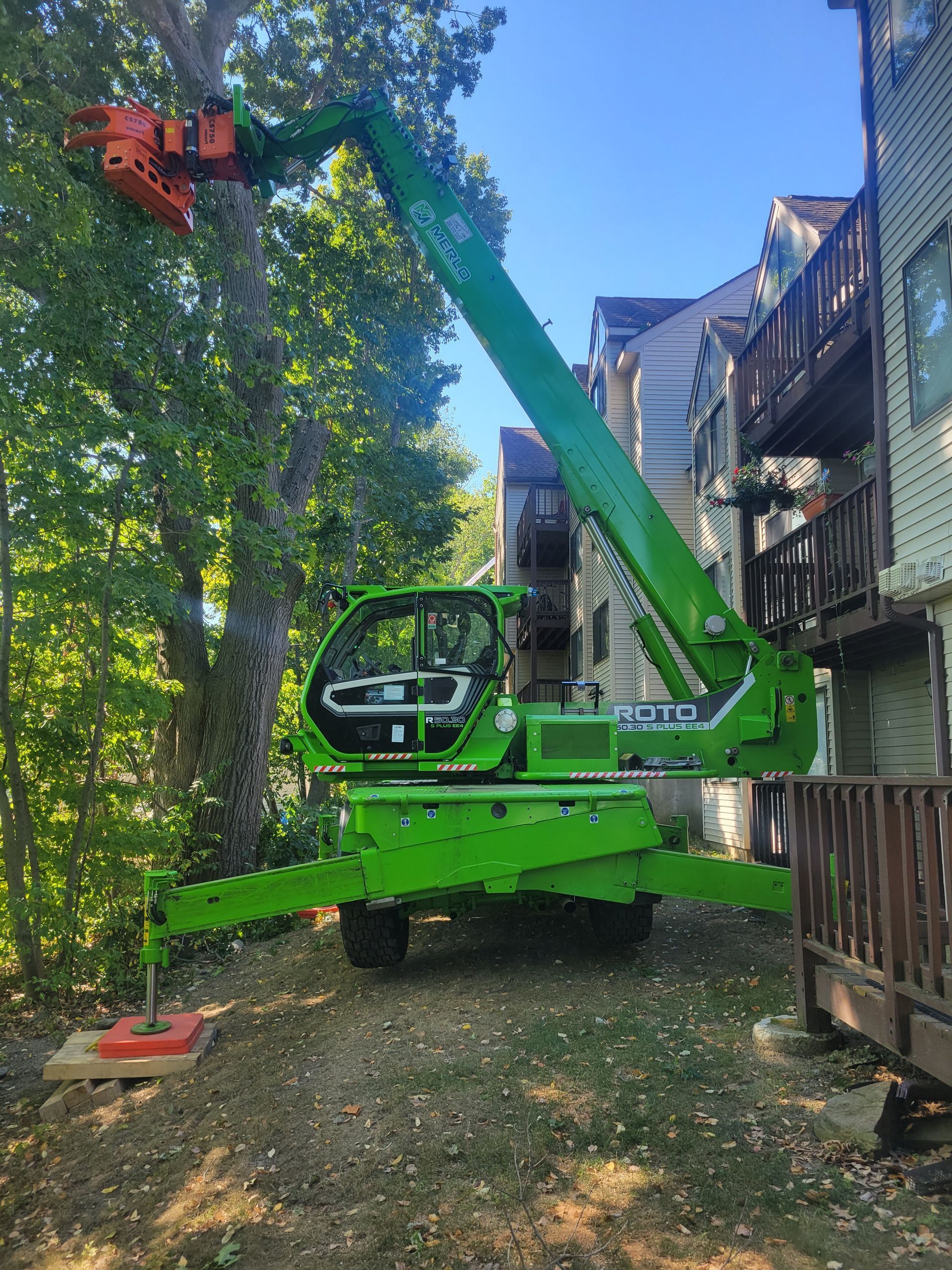 A green crane is cutting a tree in front of a building.