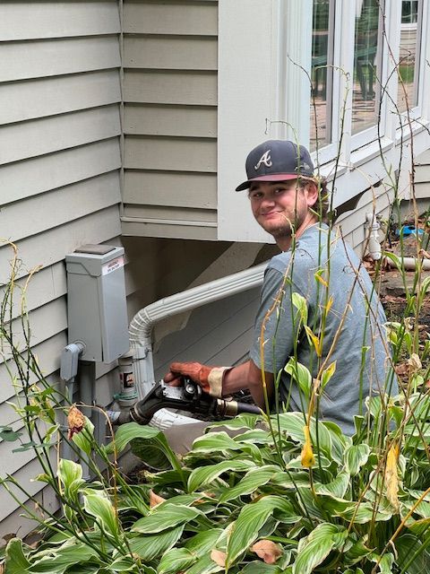 A young man in a hat smiles while working on an outdoor utility box near a house, surrounded by plants.