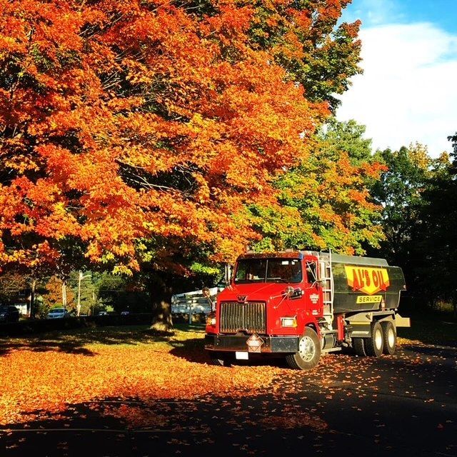 Red tanker truck parked beneath vibrant orange autumn trees.