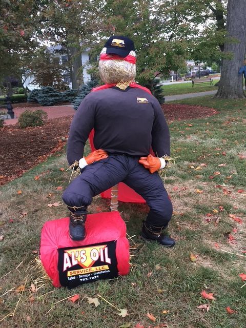 Scarecrow in work clothes posed on a red oil container with a logo; outdoors.