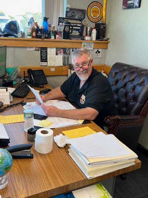 Man in office chair at desk, holding papers, smiling. Desk has papers, phone, toilet paper, and a globe.