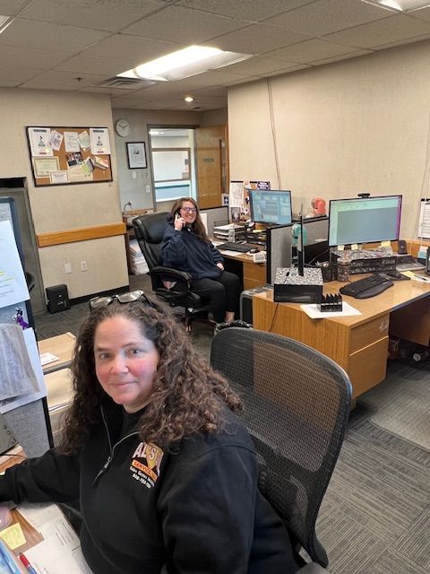 Two women at desks in an office. One smiles at the camera; the other talks on a phone.