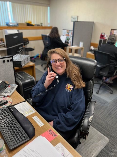 Woman in blue sweatshirt on a phone at a desk in an office setting, smiling.