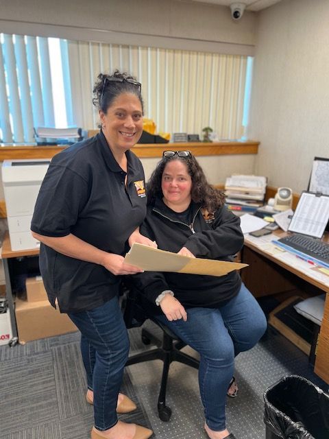 Two women in an office. One standing, holding papers, other seated. Both are smiling.