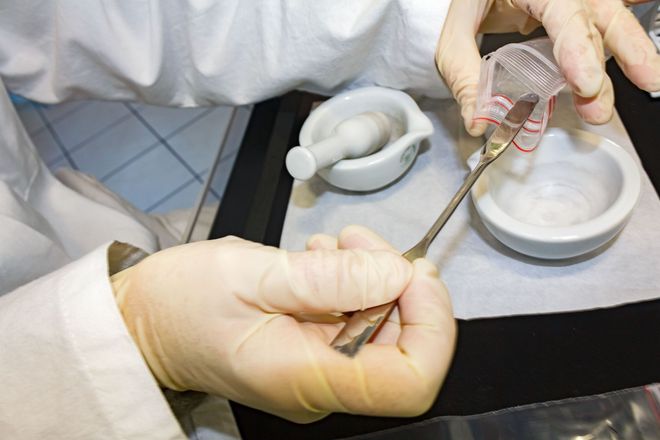 Gloved hands using a spatula to transfer substance from a beaker into a mortar and pestle on a lab table.
