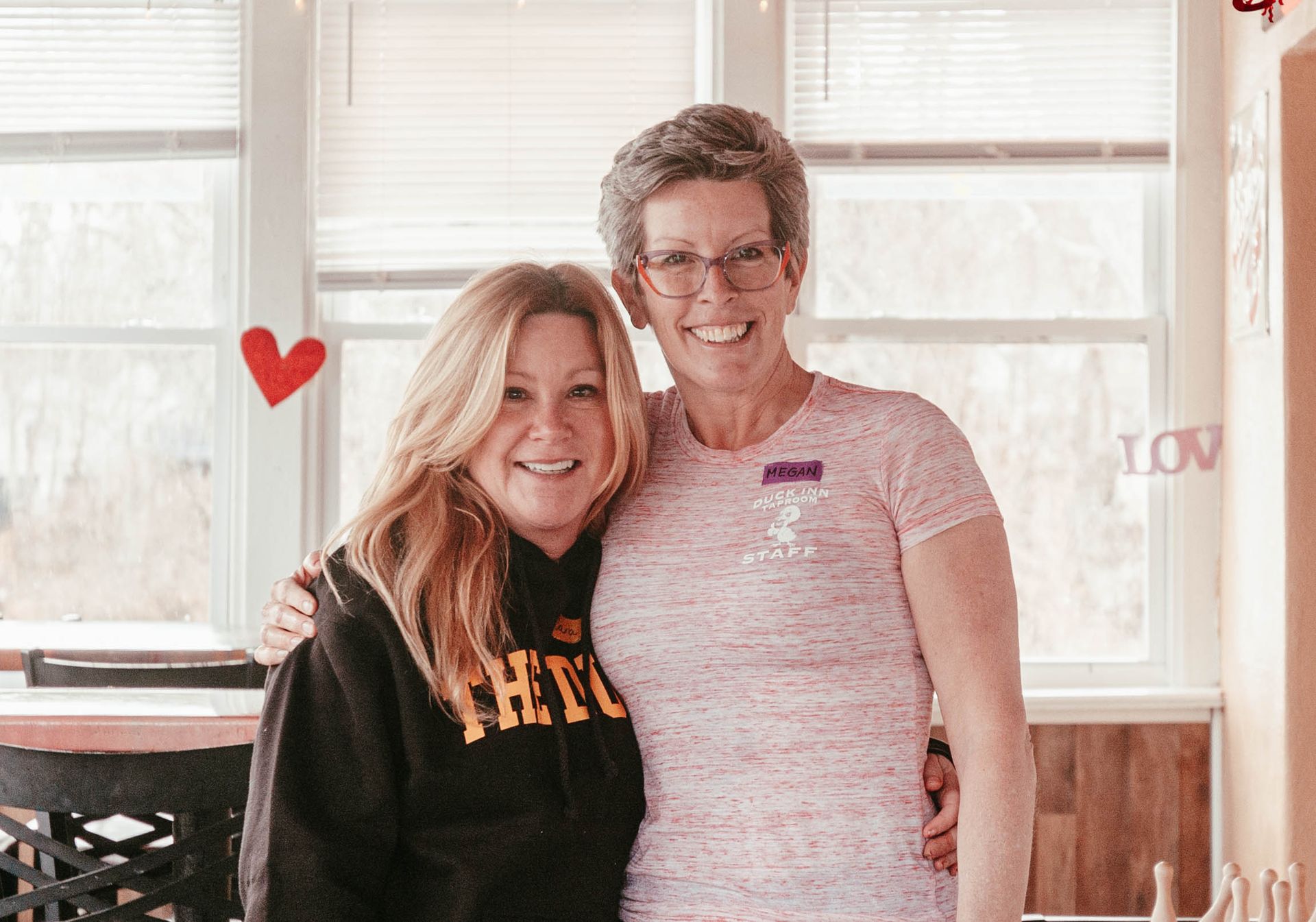 Two women are posing for a picture together in front of a window.