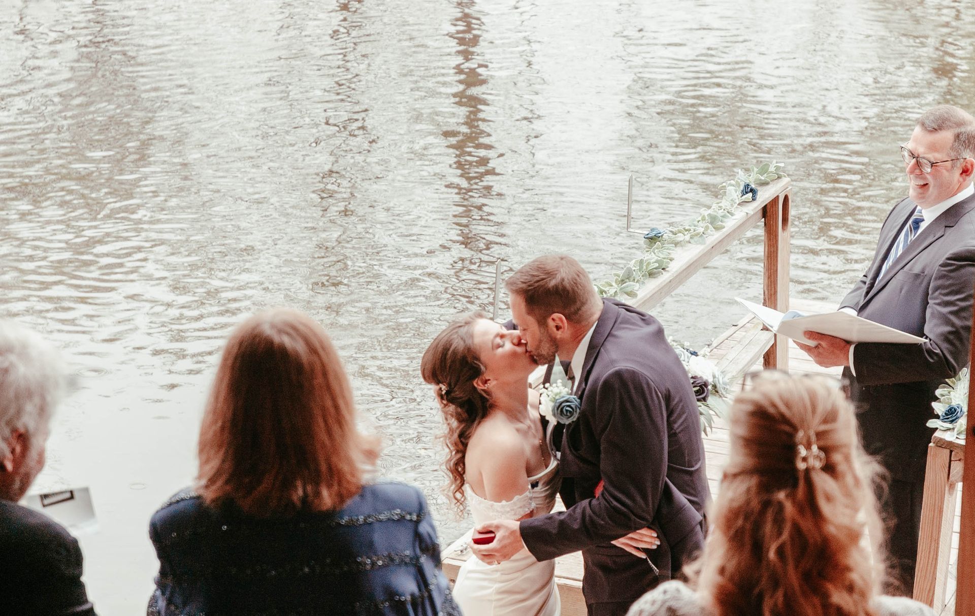 A bride and groom kissing during their wedding ceremony in front of a lake.