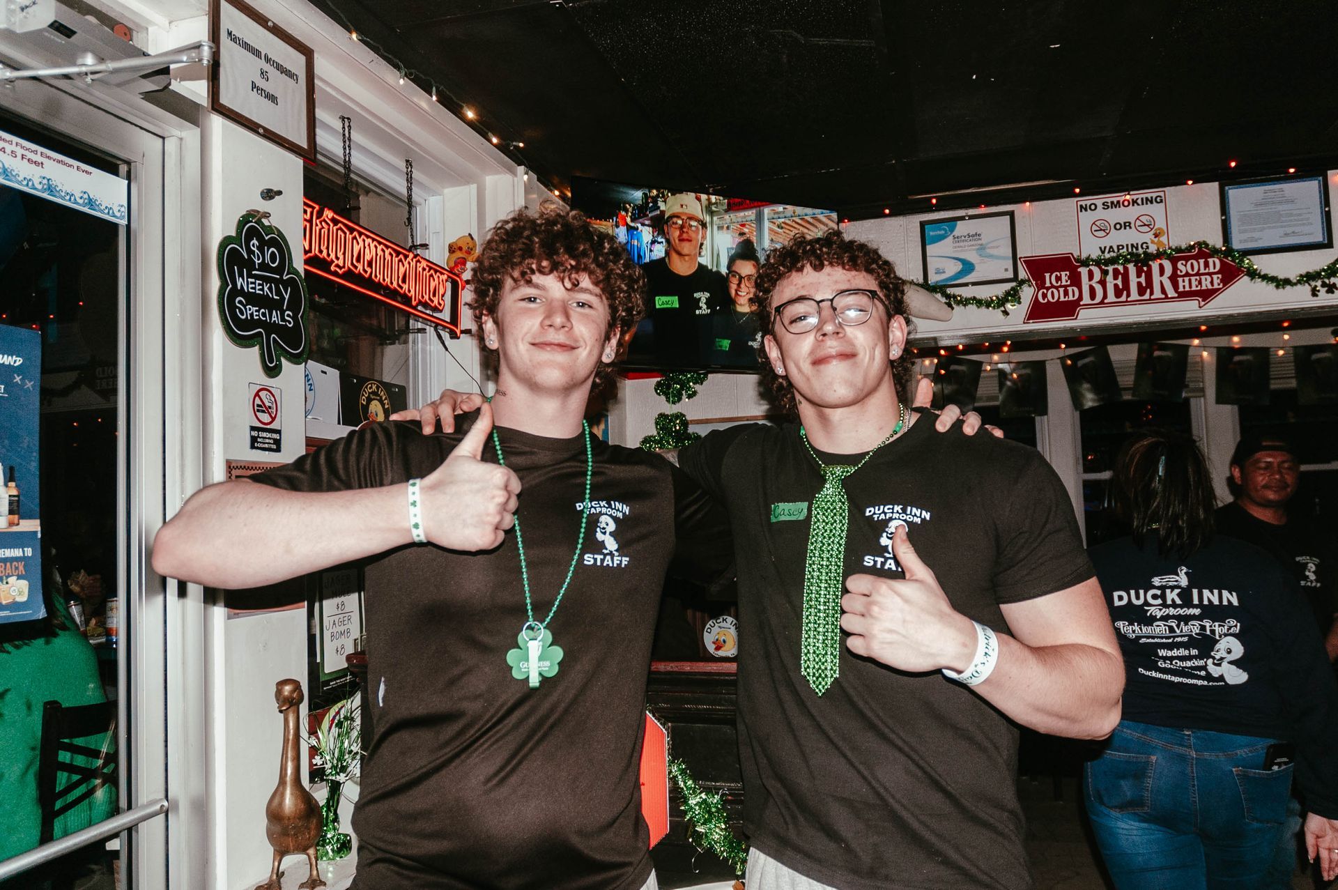 Two young men are standing next to each other in a bar giving a thumbs up.