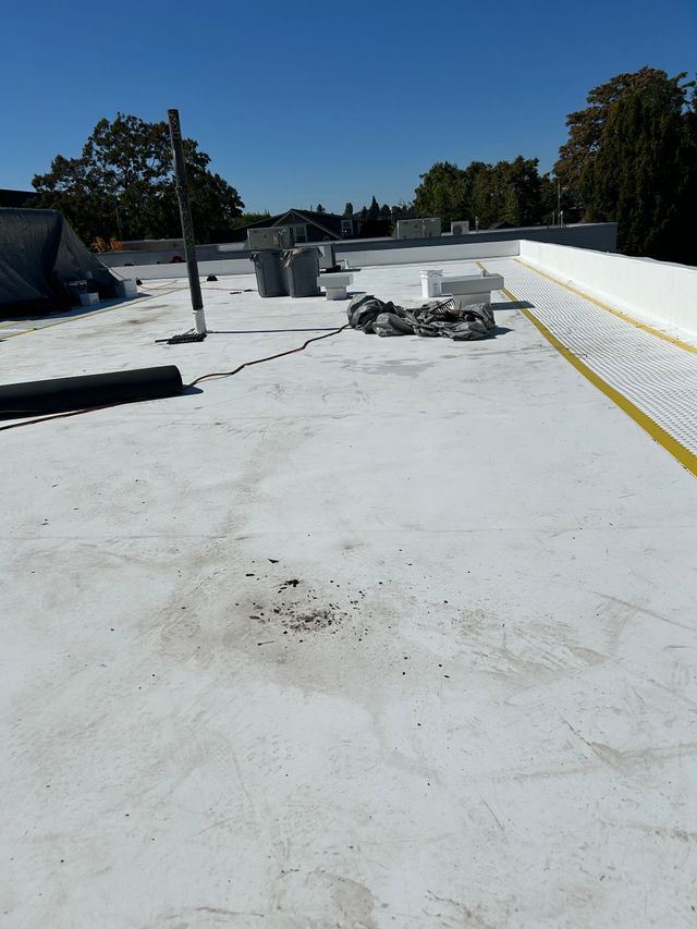 Flat white rooftop with vents, low walls, scattered debris, and trees under a clear blue sky