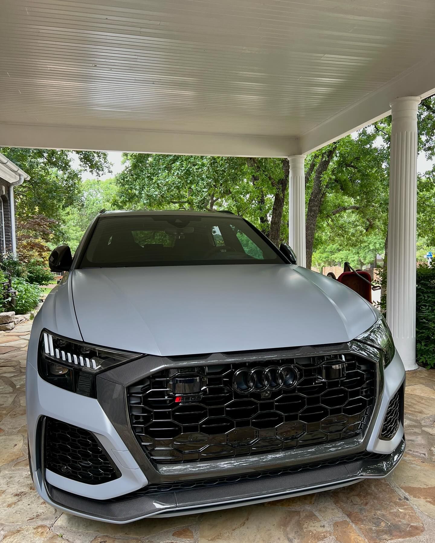 Gray Audi RS Q8 parked under a covered porch. Black grill, headlights, and front bumper. Trees in the background.