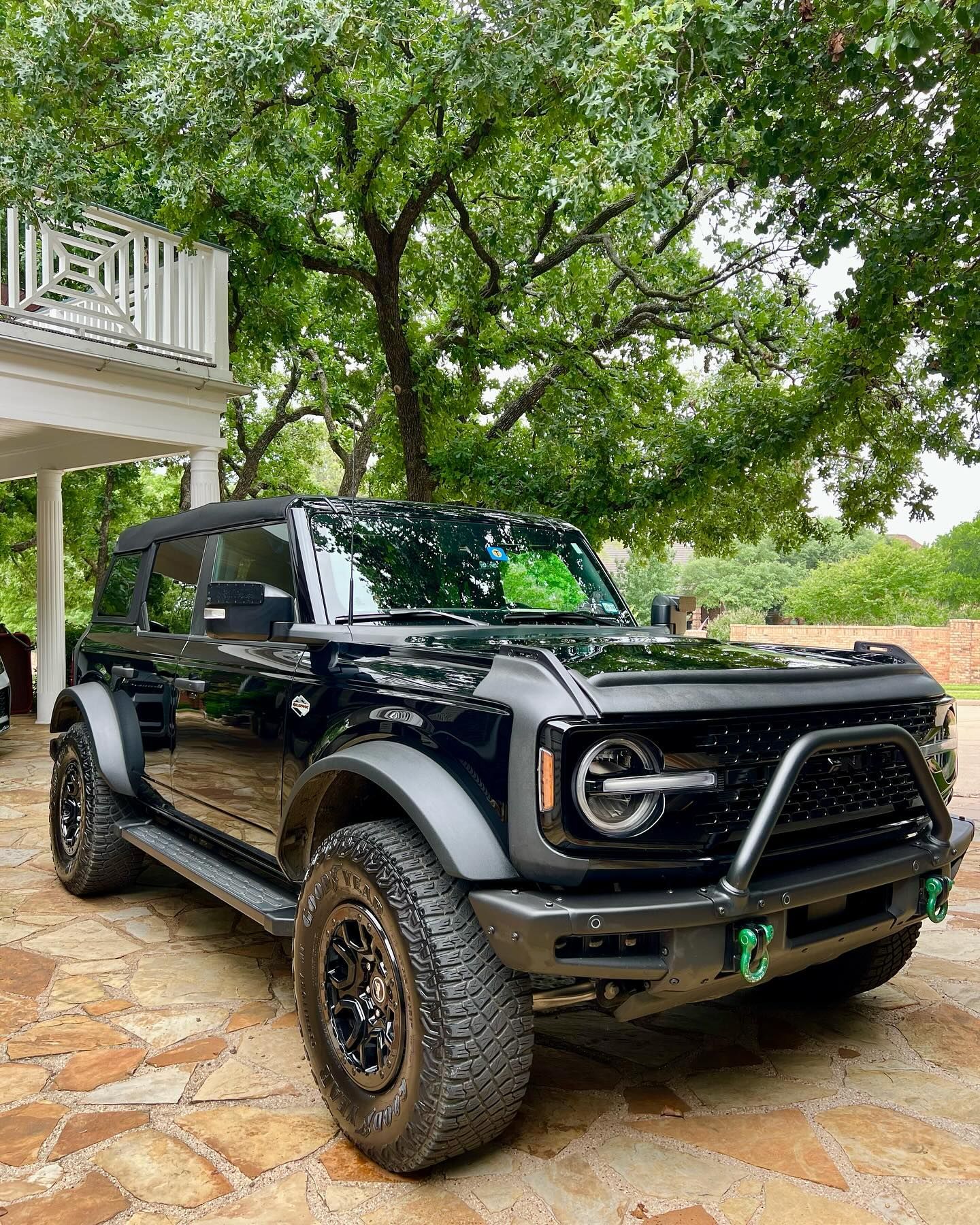 Black Ford Bronco SUV parked on a stone patio in front of a house, under a tree.
