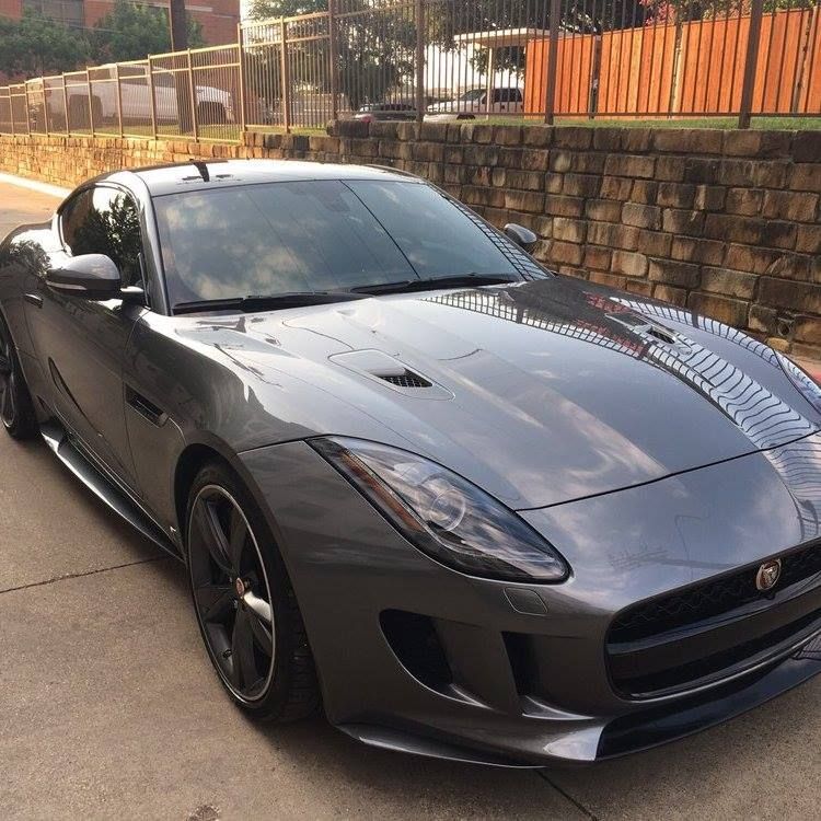 Gray Jaguar F-Type sports car parked on a paved area with a brick wall in the background.