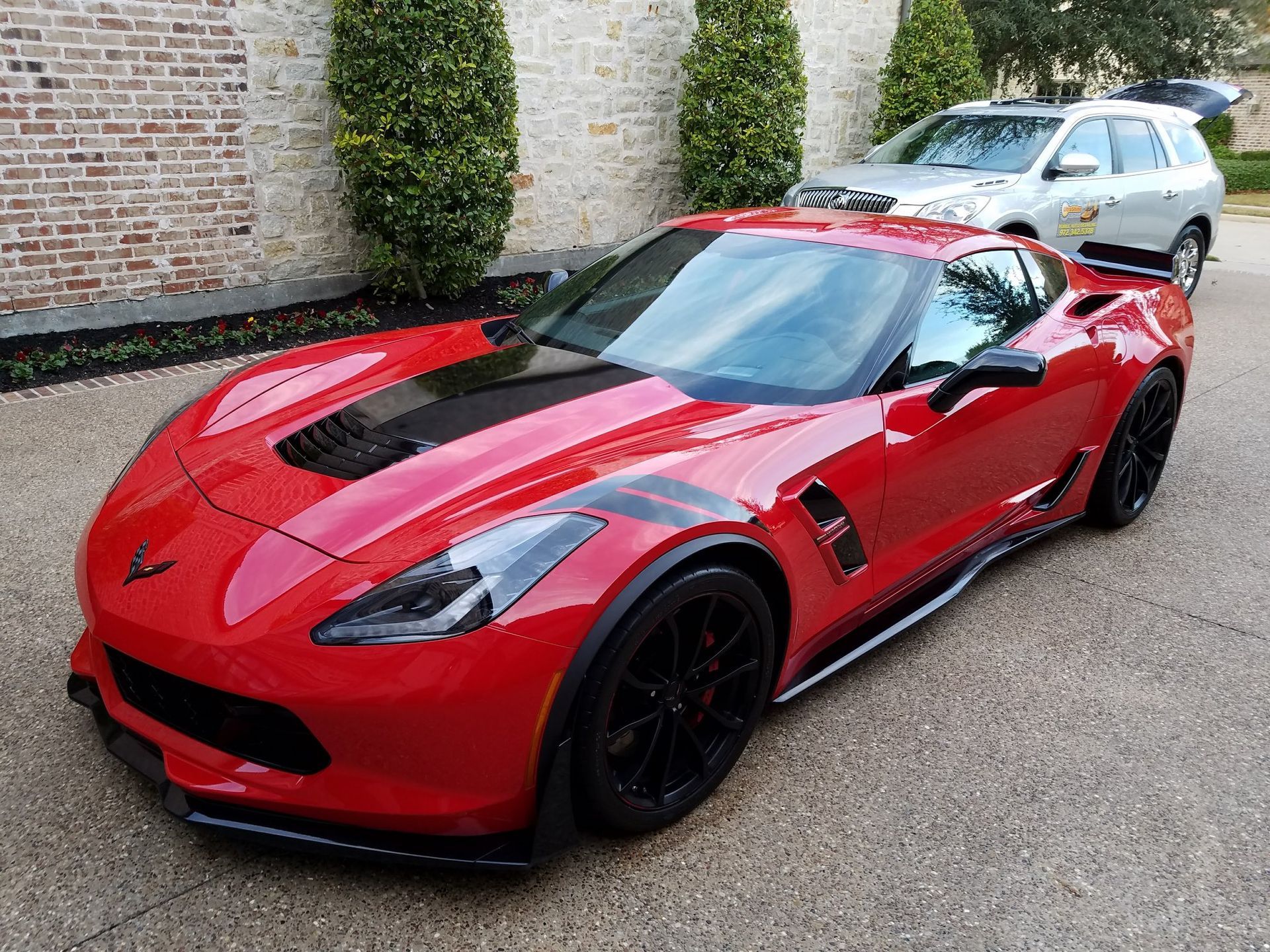 Red sports car with black accents parked on a paved driveway, a silver SUV in the background.
