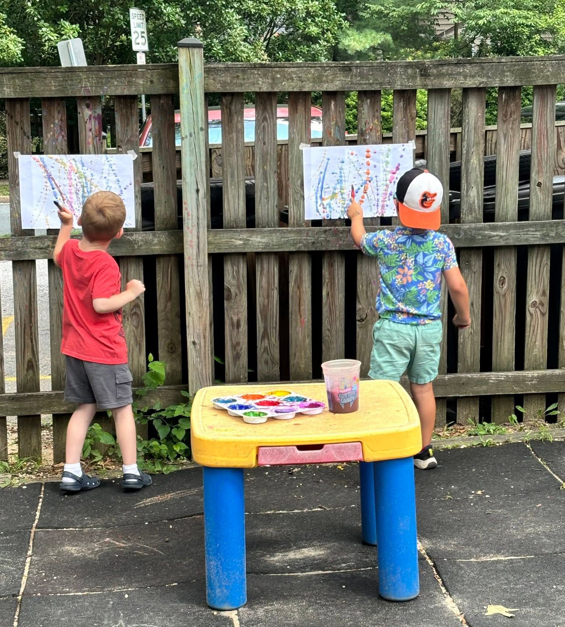 Two children painting at an outdoor easel beside a small table in a backyard
