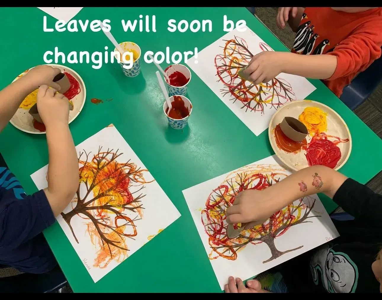 Children paint autumn trees with red, yellow, and orange leaves at a classroom table.