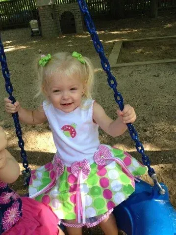 Smiling child on a blue swing, wearing a white top and bright pink-green skirt.
