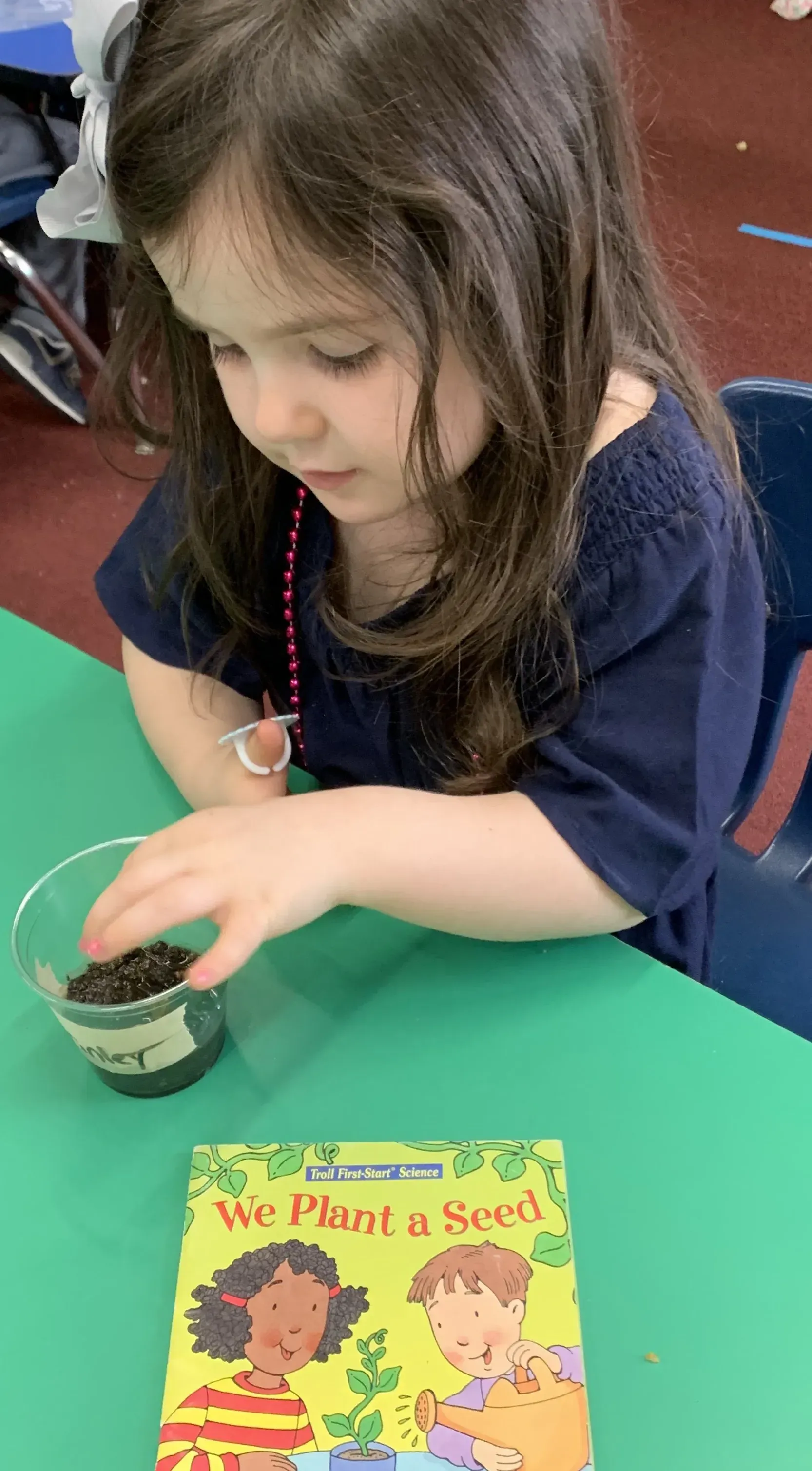 Child with long brown hair reaching into a small cup on a table beside a colorful book.