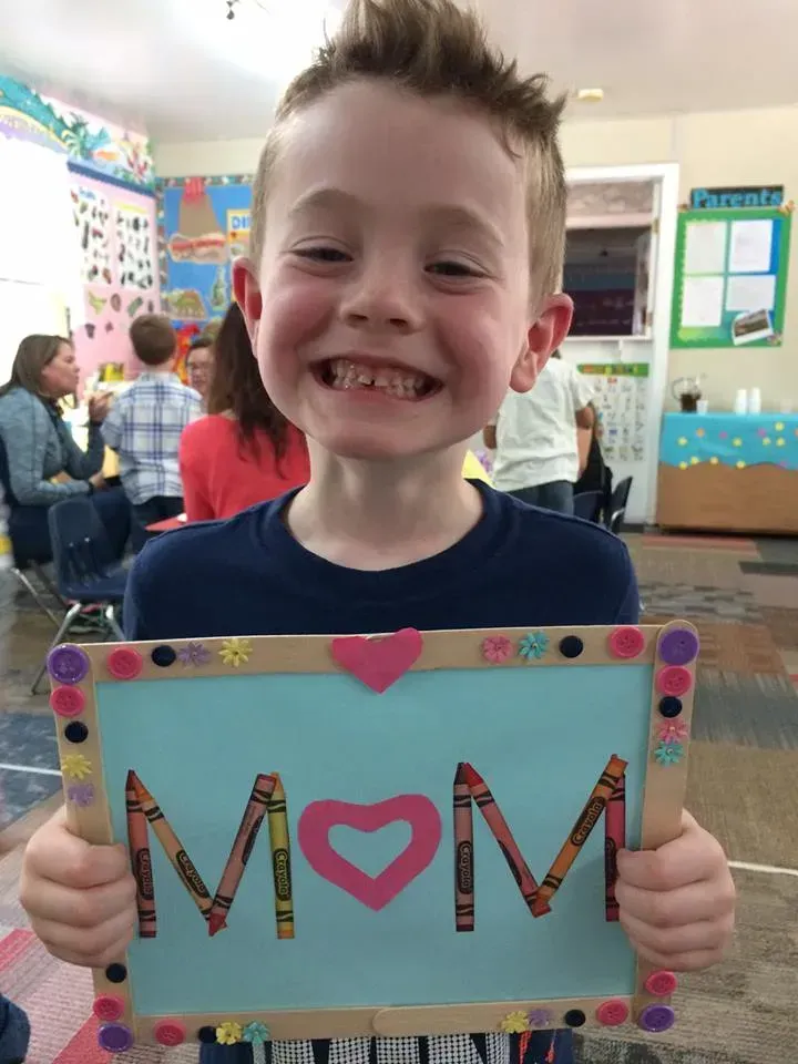 Smiling child holding a colorful Mother’s Day card that says “MOM” in a classroom.