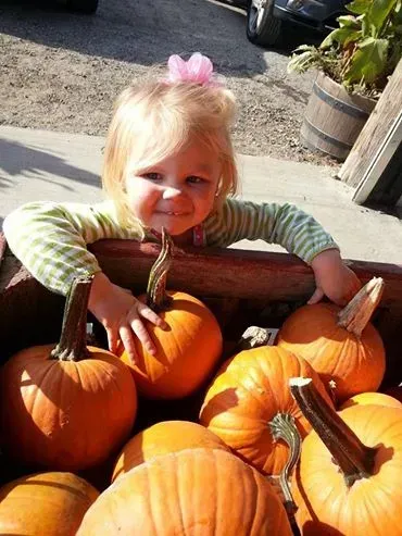 Child peeking over a pile of orange pumpkins outdoors, wearing a pink bow and striped shirt