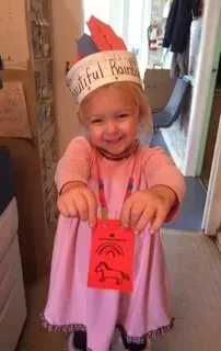 Child wearing a paper crown and pink dress smiles while holding a bright pink card.