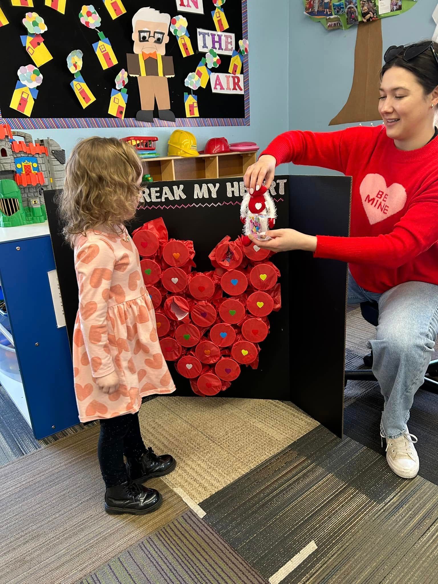 Teacher handing a red craft to a child in a classroom decorated with heart art