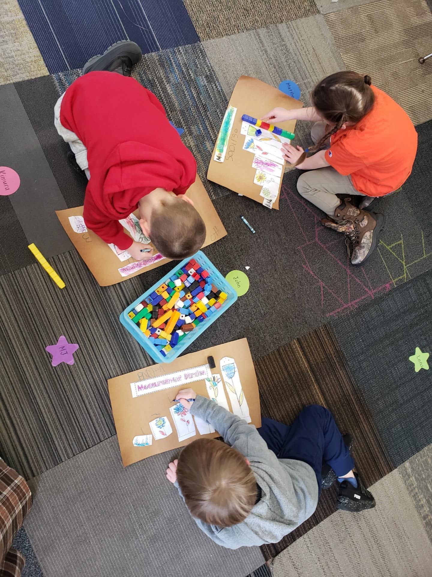 Three children sitting on carpet, drawing and playing with colorful toys and paper boards.