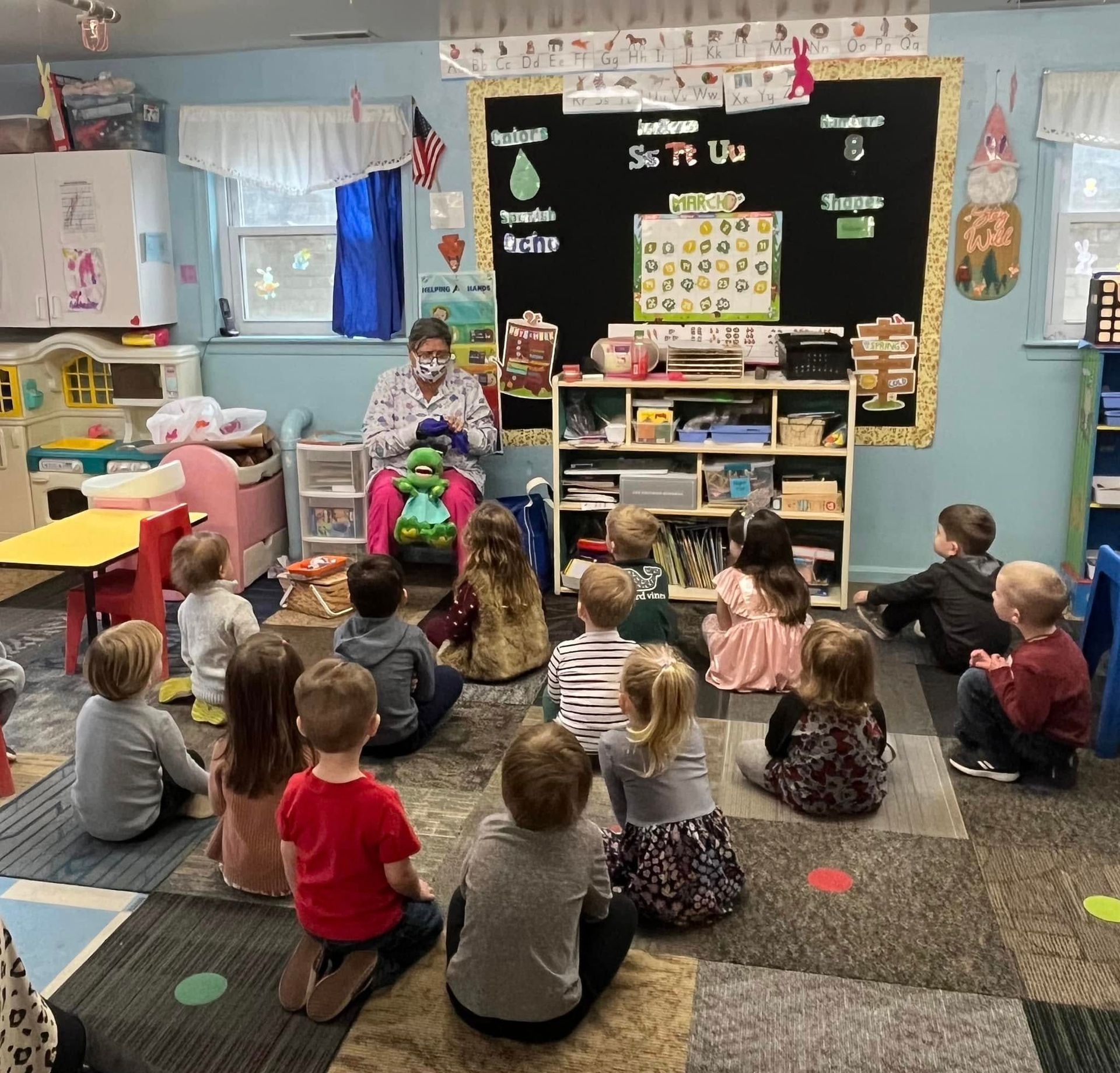 Preschool class seated on the floor listening to a teacher in a colorful classroom near a blackboard.