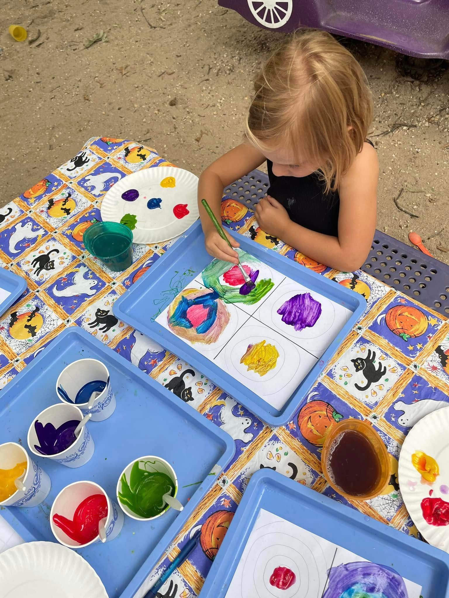 Child painting colorful shapes at an outdoor art table with paint cups and papers