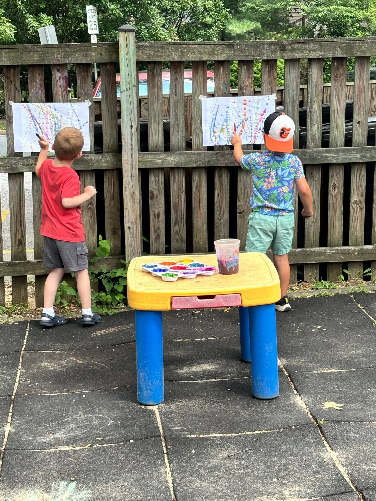 Two children painting papers on a wooden fence beside a small table outdoors