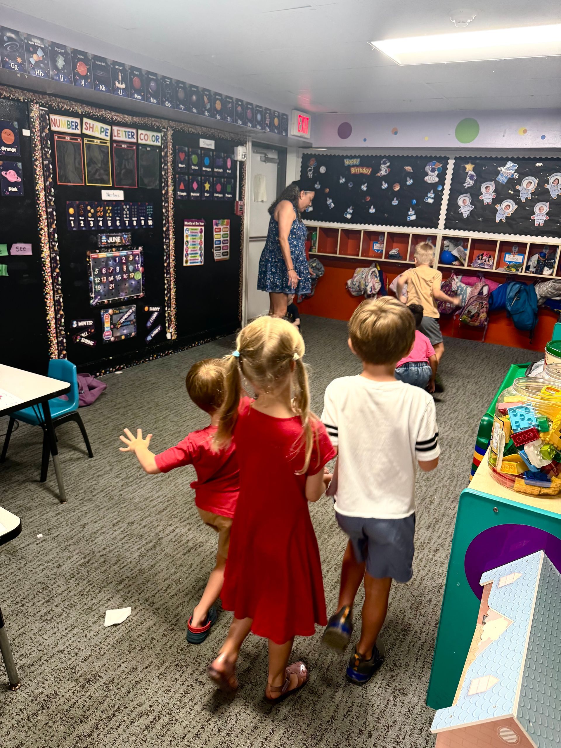 Children in a classroom playing near cubbies, with a teacher standing at the front.