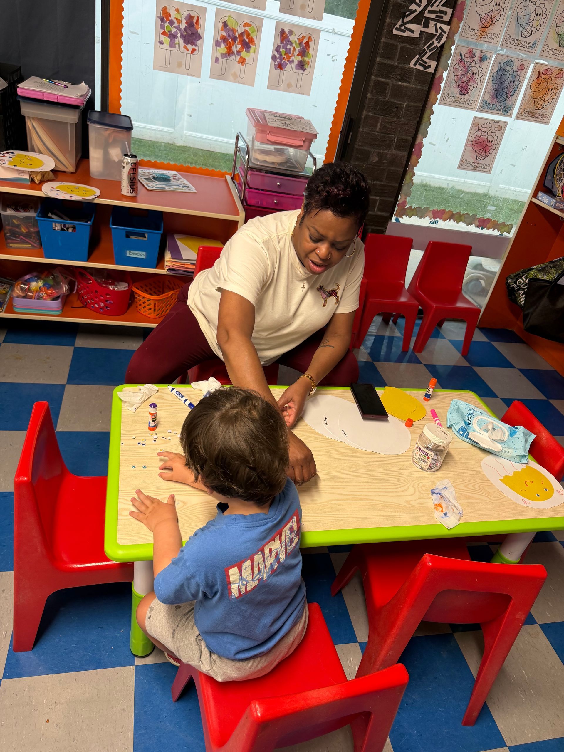 Teacher and child painting together at a small red table in a colorful classroom.