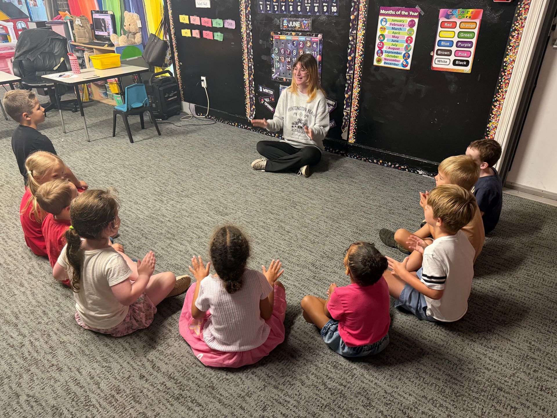 Children sit in a circle on a carpet around a teacher in a classroom.