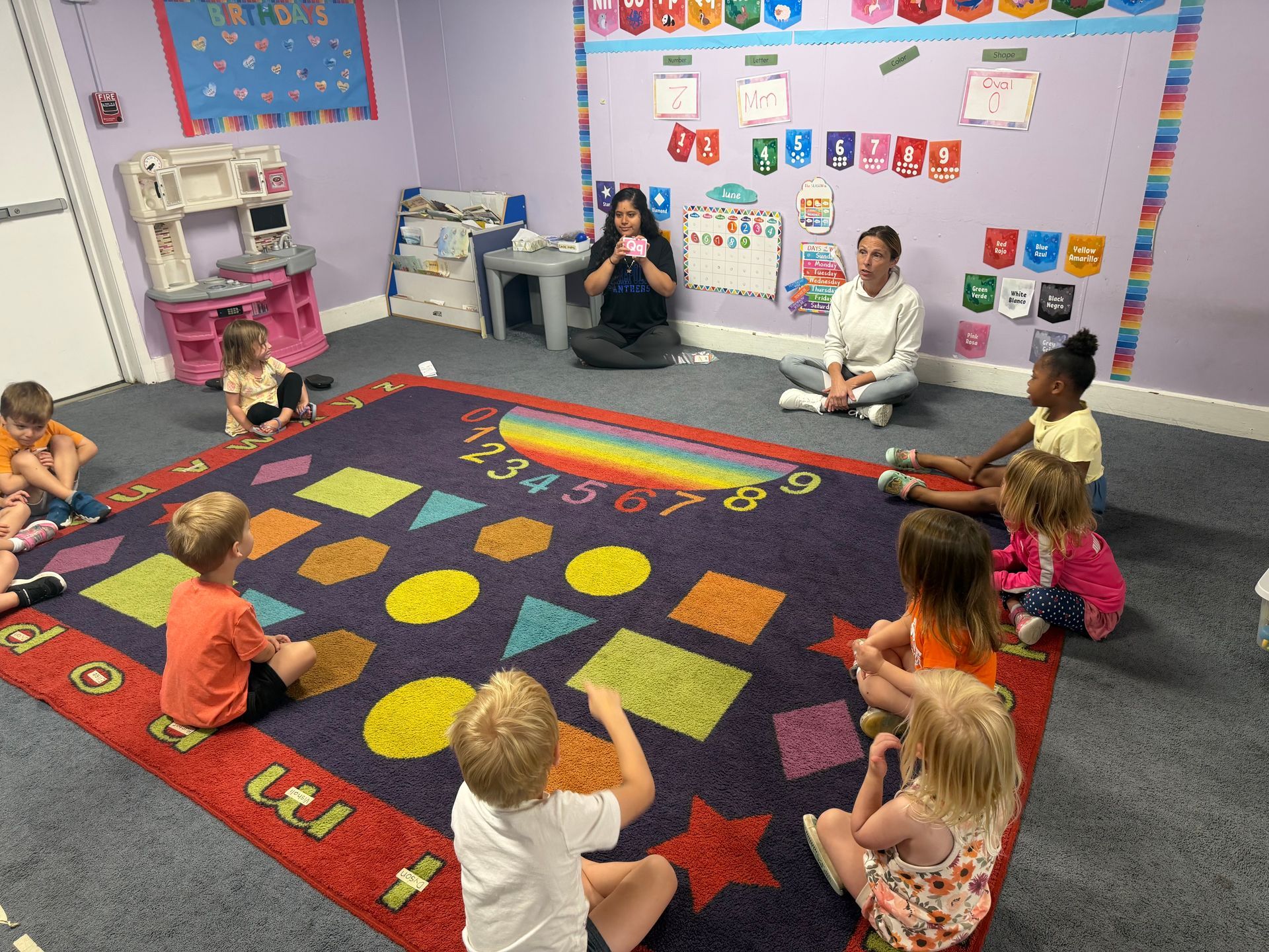 Children sit in a colorful classroom circle on a patterned rug, facing two seated teachers.