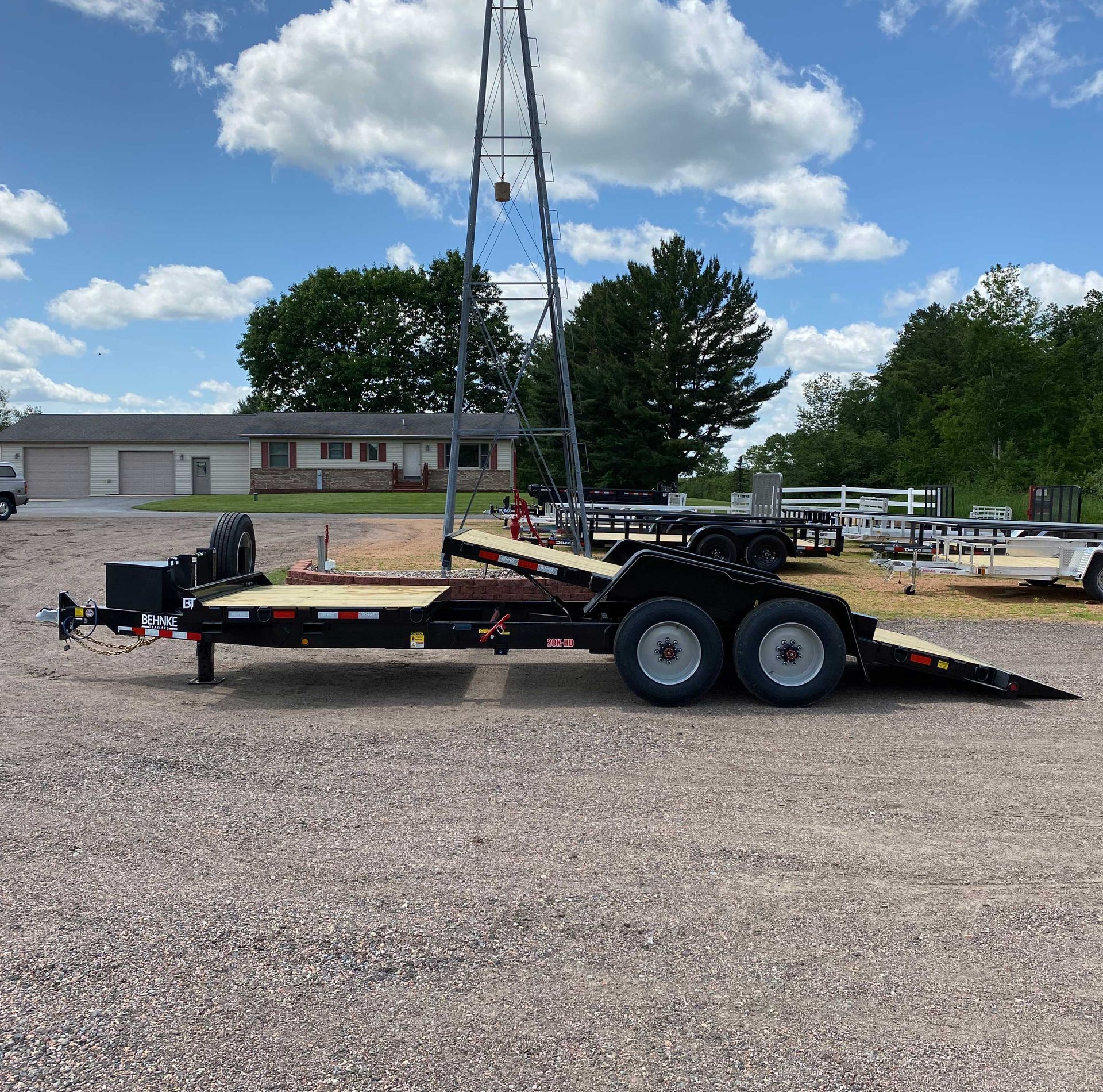 Black trailer with wooden deck on gravel, under a blue sky.