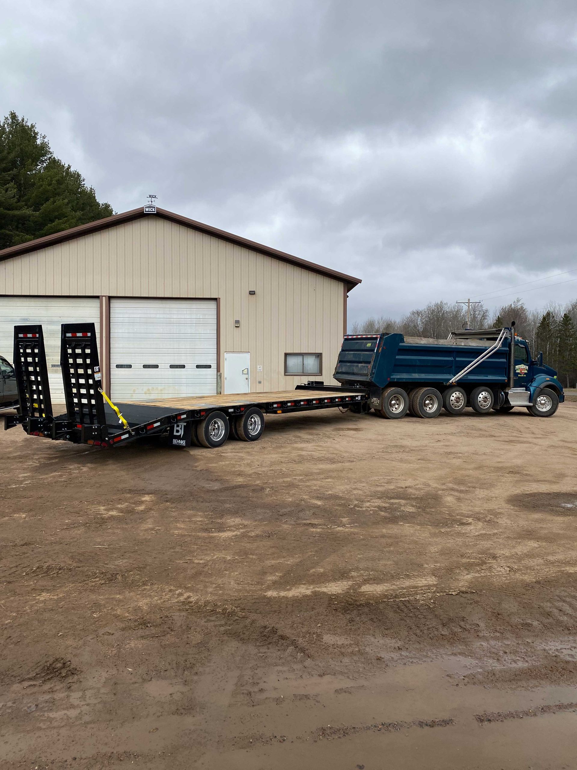 Blue dump truck with trailer parked outside a beige building on a cloudy day.