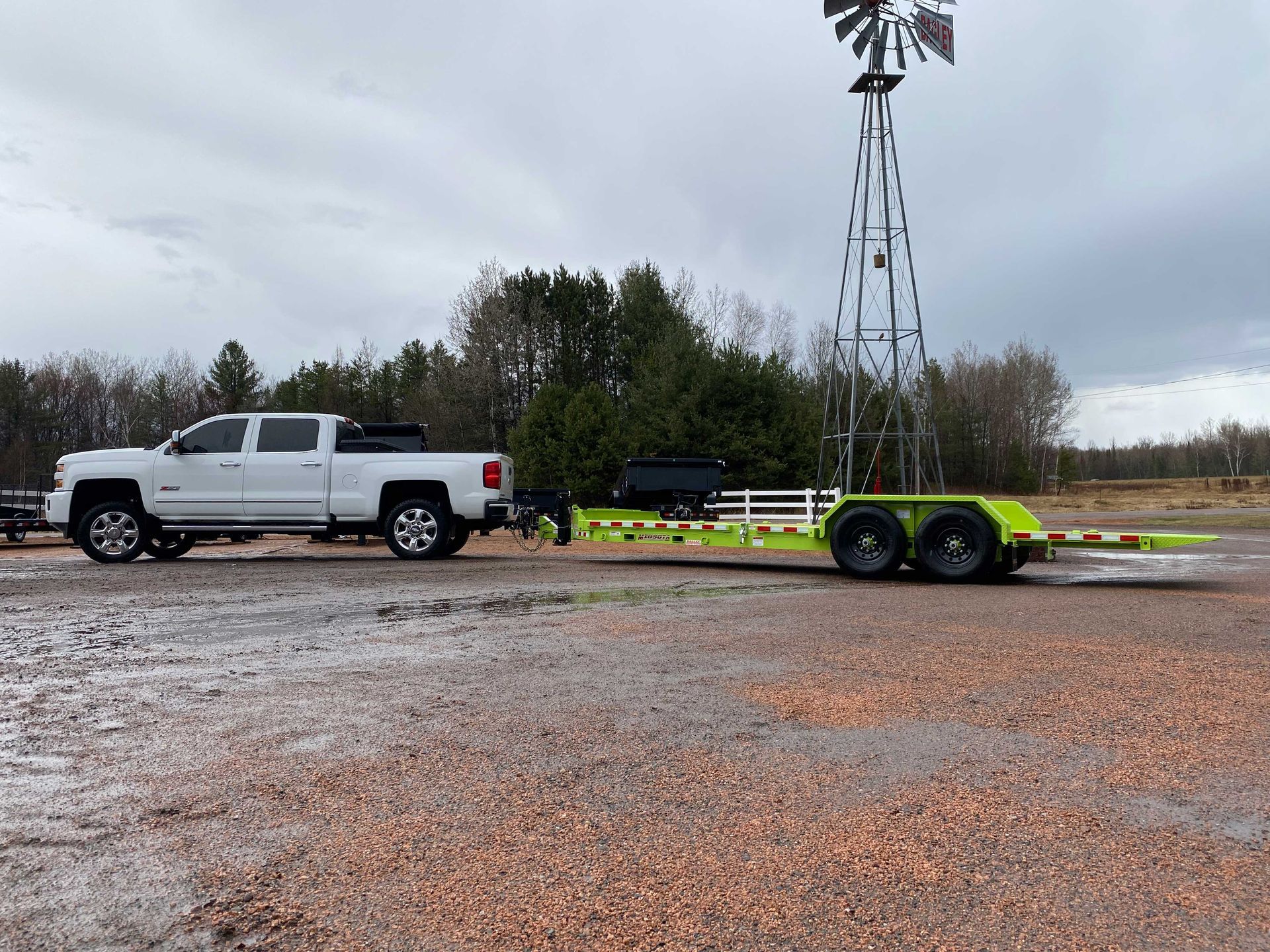 White truck towing a bright green trailer, near a windmill in a rural area under an overcast sky.